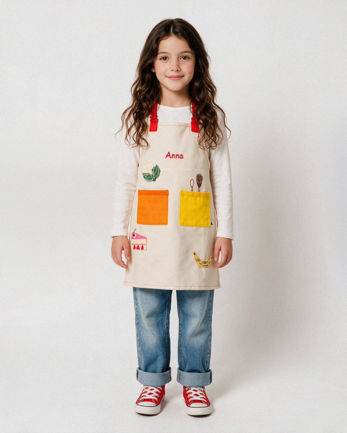 Petite fille portant un tablier de cuisine enfant ADADE beige, personnalisé avec le prénom Anna brodé en rouge et des patchs thermocollants colorés représentant des aliments