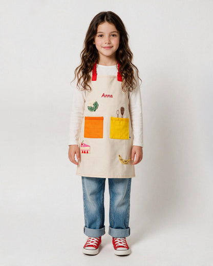 Petite fille portant un tablier de cuisine enfant ADADE beige, personnalisé avec le prénom Anna brodé en rouge et des patchs thermocollants colorés représentant des aliments